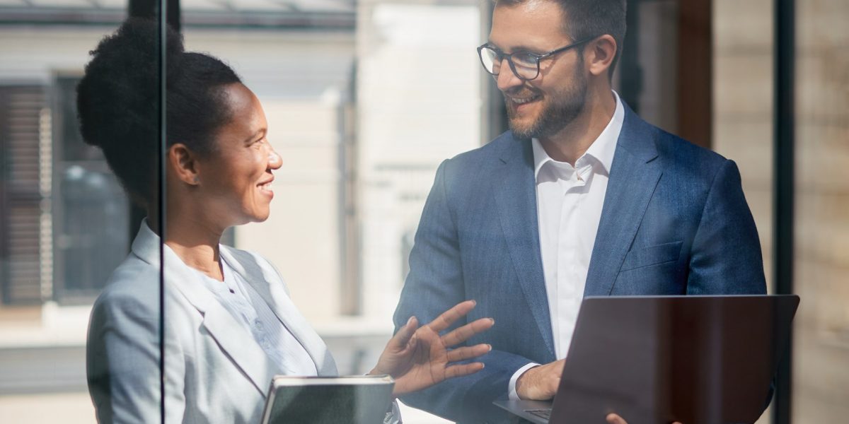 Happy businessman using laptop while communicating with African American female colleague at corporate office. The view is through the glass.